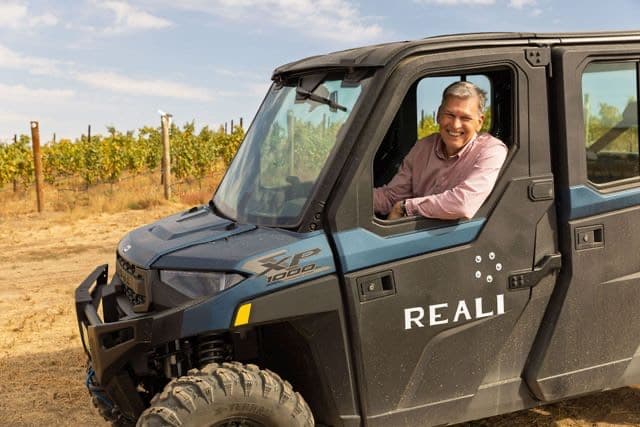 a man leans out of the window of a UTV with the word REALI on the door and there are grapevines in the background